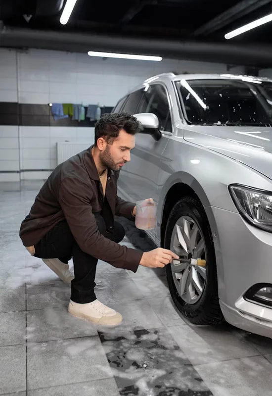 Man crouching next to a silver car, cleaning the wheel rim with a brush while holding a cup of cleaning solution.