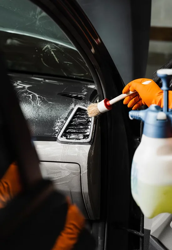 Person wearing orange gloves using a detailing brush to clean a car’s air vent, with cleaning foam and a spray bottle visible.