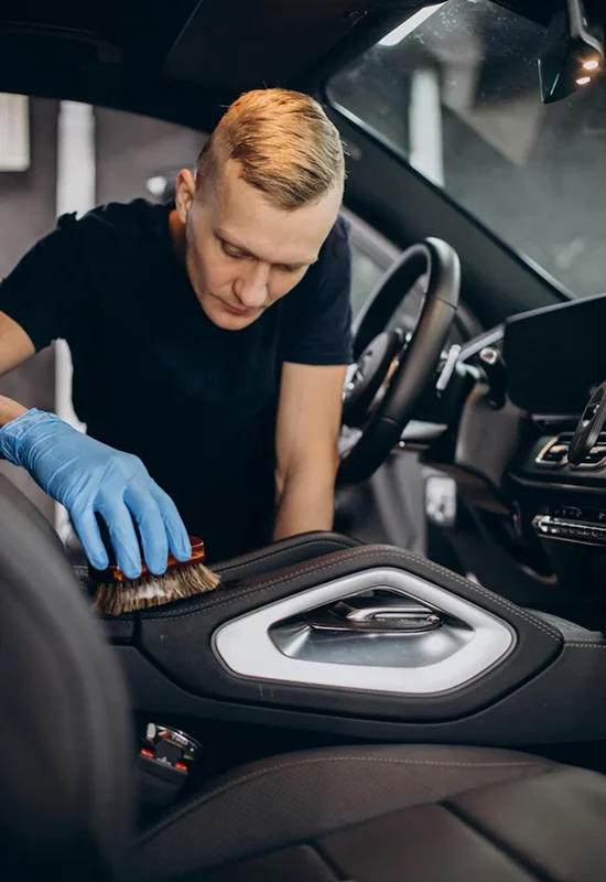 Man wearing blue gloves cleaning a car’s interior door panel with a detailing brush.