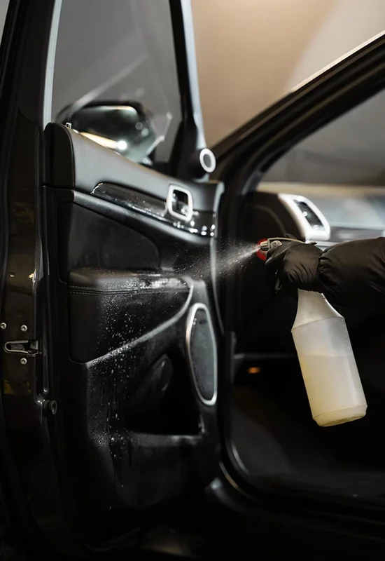 Person wearing black gloves spraying cleaner onto the interior door panel of a car.