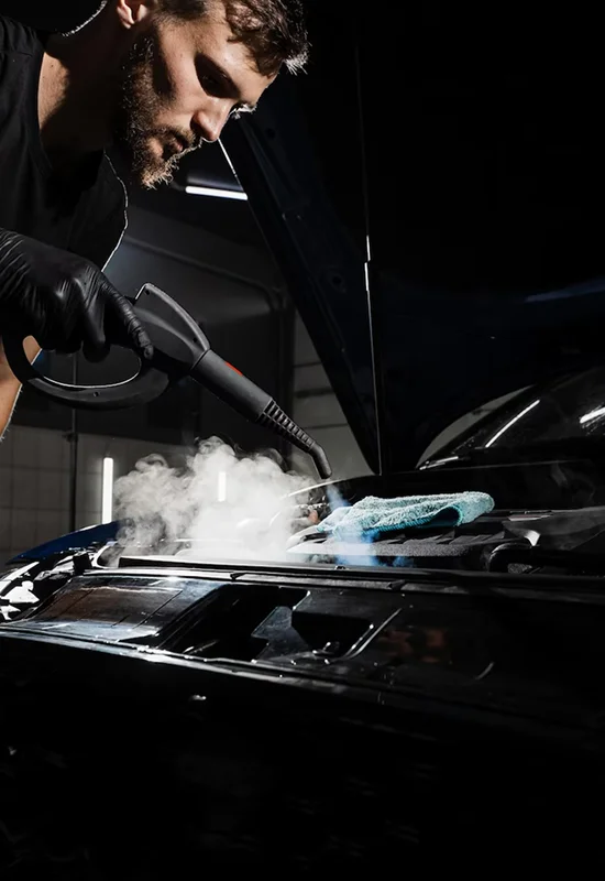 Man using a steam cleaning tool under a car’s hood with steam rising and a microfiber cloth nearby.