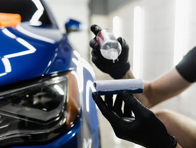 Person wearing black gloves applying liquid from a bottle onto an applicator pad in front of a blue car’s headlight.