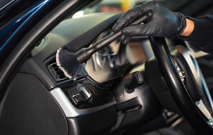 Person wearing black gloves using a detailing brush to clean the air vent and dashboard area of a car interior.