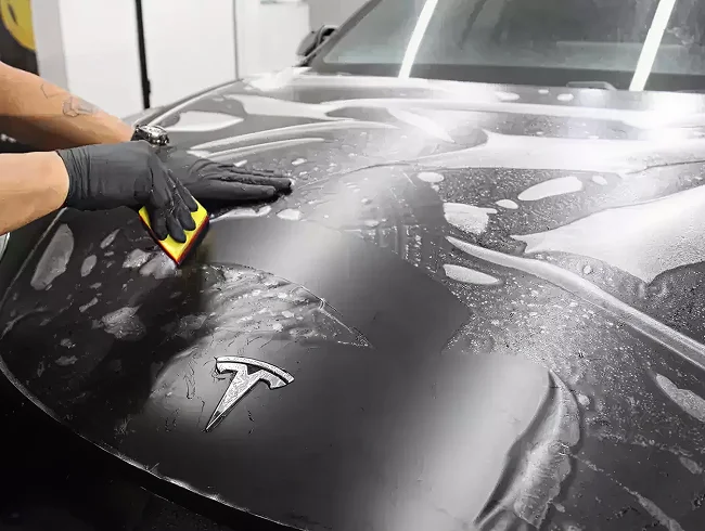 Person wearing black gloves applying a protective film to the hood of a black Tesla, using a yellow squeegee to smooth out water and bubbles.