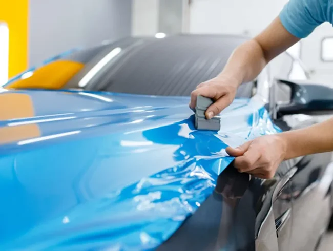 Person applying a glossy blue vinyl wrap to a car’s hood using a squeegee tool to smooth the surface.