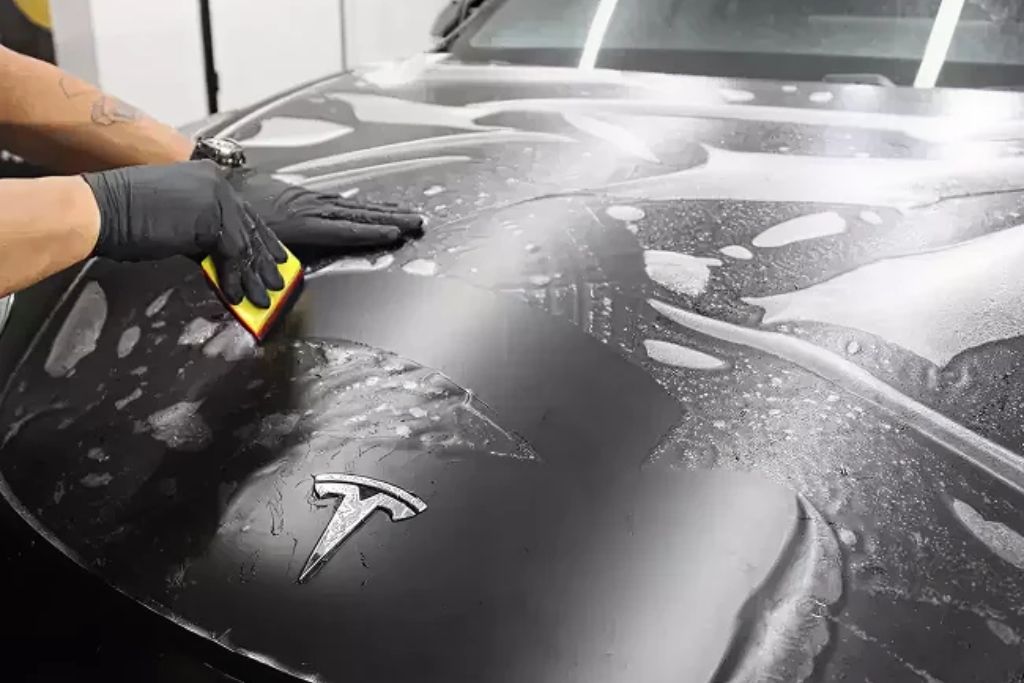Technician applying paint protection film to the hood of a black Tesla using a squeegee, with water and solution visible during installation. This image represents professional service and contributes to understanding paint protection film cost based on vehicle size and coverage.