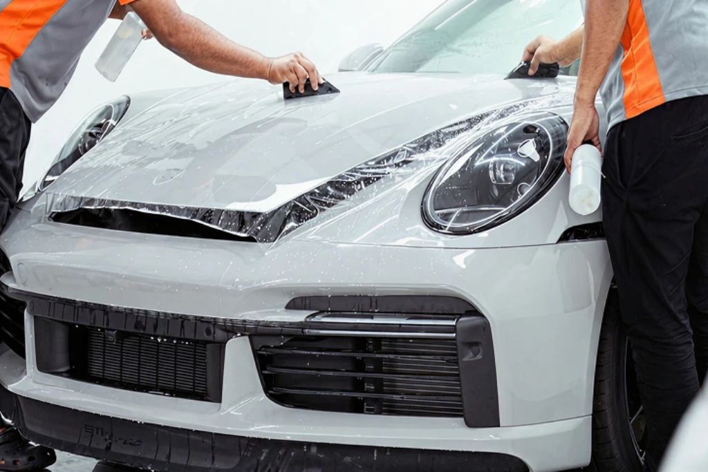 Two technicians professionally install paint protection film on the front of a white sports car, carefully smoothing the clear layer onto the hood and fenders. This image demonstrates the expert process behind what is paint protection film and the importance of proper application.