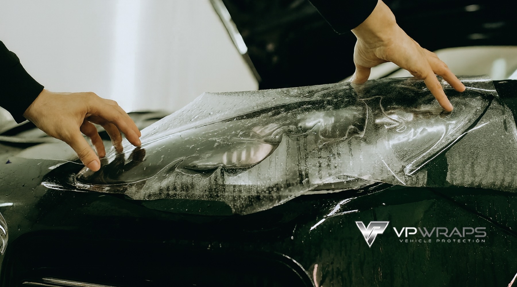 Close-up of a technician's hands applying a transparent paint protection film over a wet car hood and headlight, showcasing the detailing process. The VP Wraps logo is visible in the lower corner, indicating a professional service based in Magnolia, TX.