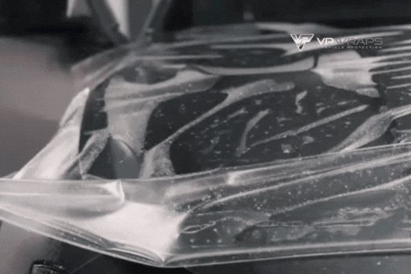 A sheet of paint protection film is carefully laid over the hood of a car at VP Wraps during installation, with water and slip solution visible beneath the film for proper alignment and adhesion.