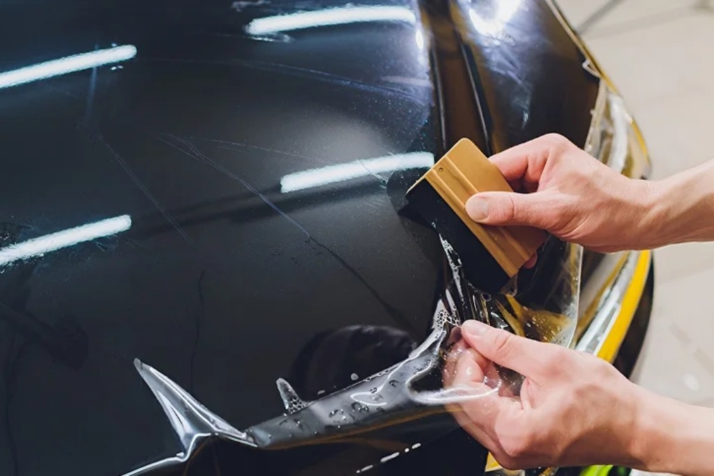 Close-up of a technician applying PPF installation to a black car hood using a squeegee, with the clear film partially adhered and air bubbles being smoothed out for a seamless finish.