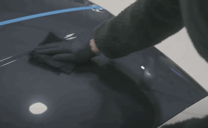 Gloved hand applies ceramic coating to half of a car hood, divided with blue tape.