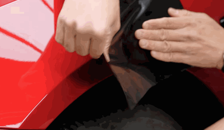 Hands applying a black vinyl wrap to the fender of a red car, smoothing the material by hand for a seamless finish.