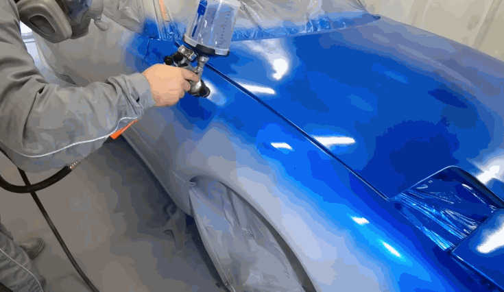 A technician in protective gear sprays glossy blue automotive paint onto a car’s fender using a paint gun inside a controlled booth.
